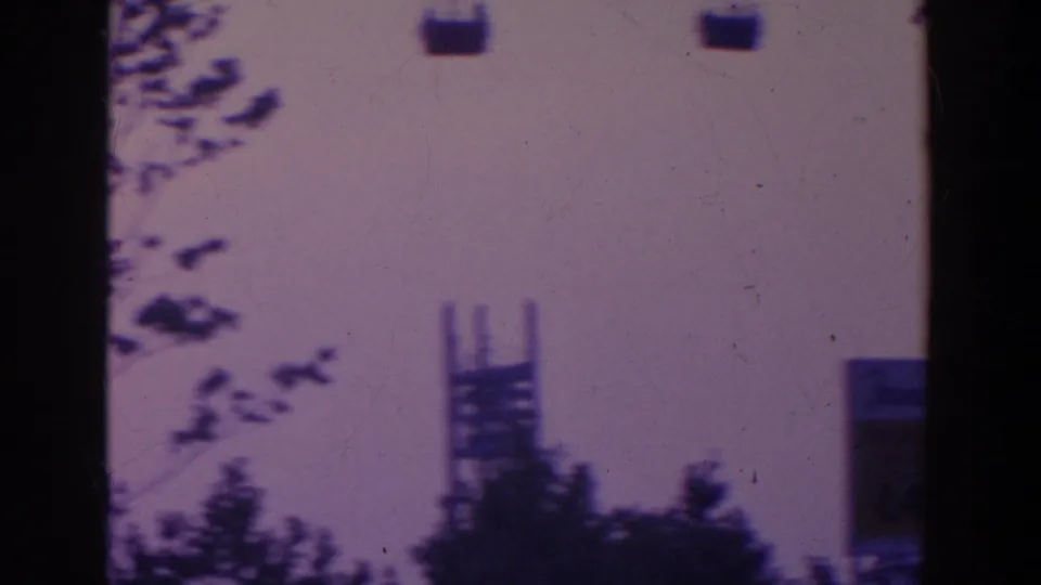 1964 8mm frame with small overhead gondolas, silhouetted trees, and a tall fairground support against a pale sky.