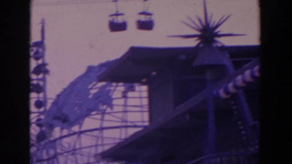1964 8mm frame showing overhead gondolas above a fairground building, with a spiky decorative form and striped pole.