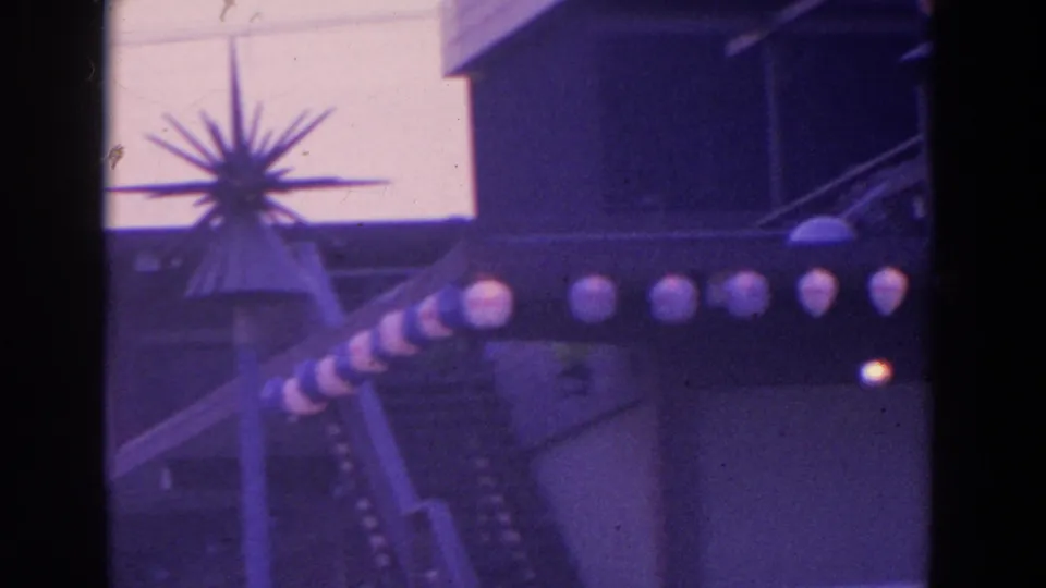 1964 8mm frame showing a fairground building edge, spiky decorative element, striped pole, and row of spherical lights.