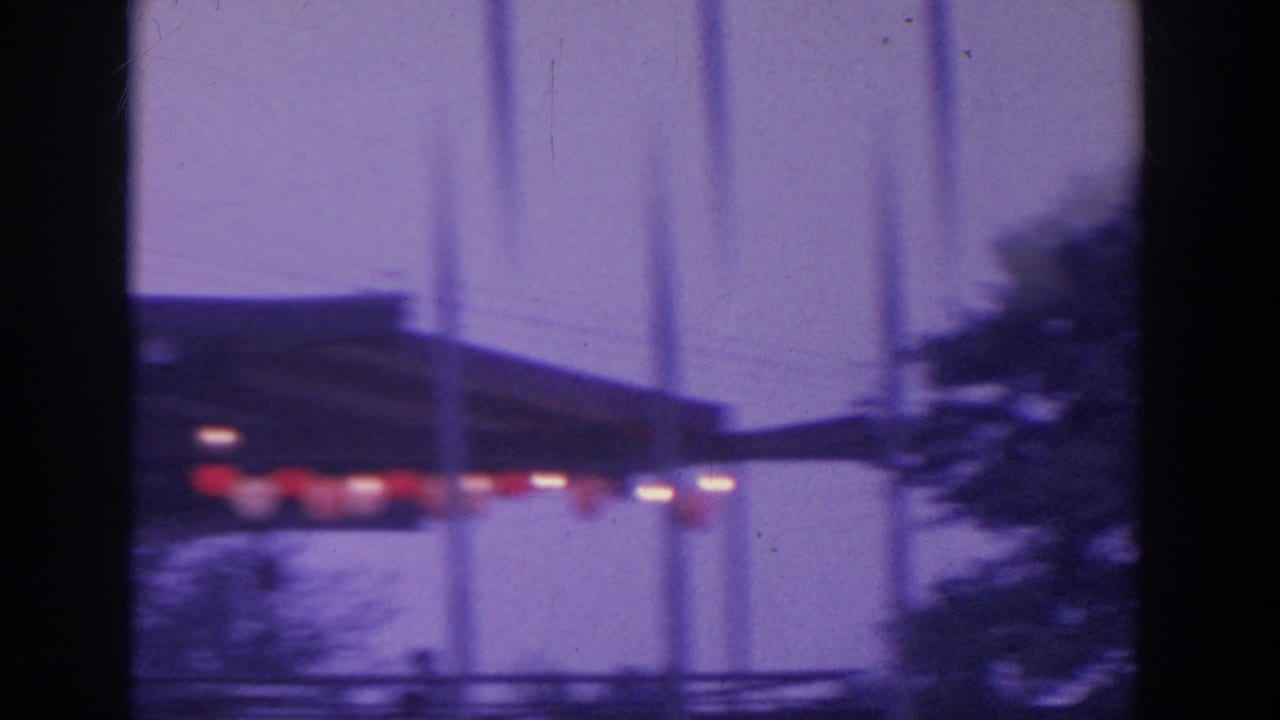 Purple-tinted 8mm frame showing a blurred structure with red lights, vertical lines, and nearby trees.