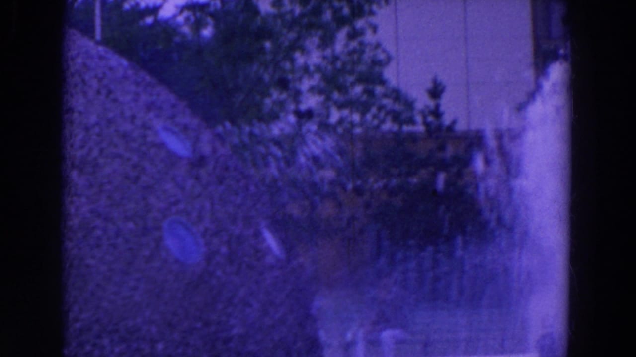 Color-shifted 8mm frame of a patterned water feature, with jets emerging from a textured slope near trees and a pale facade.