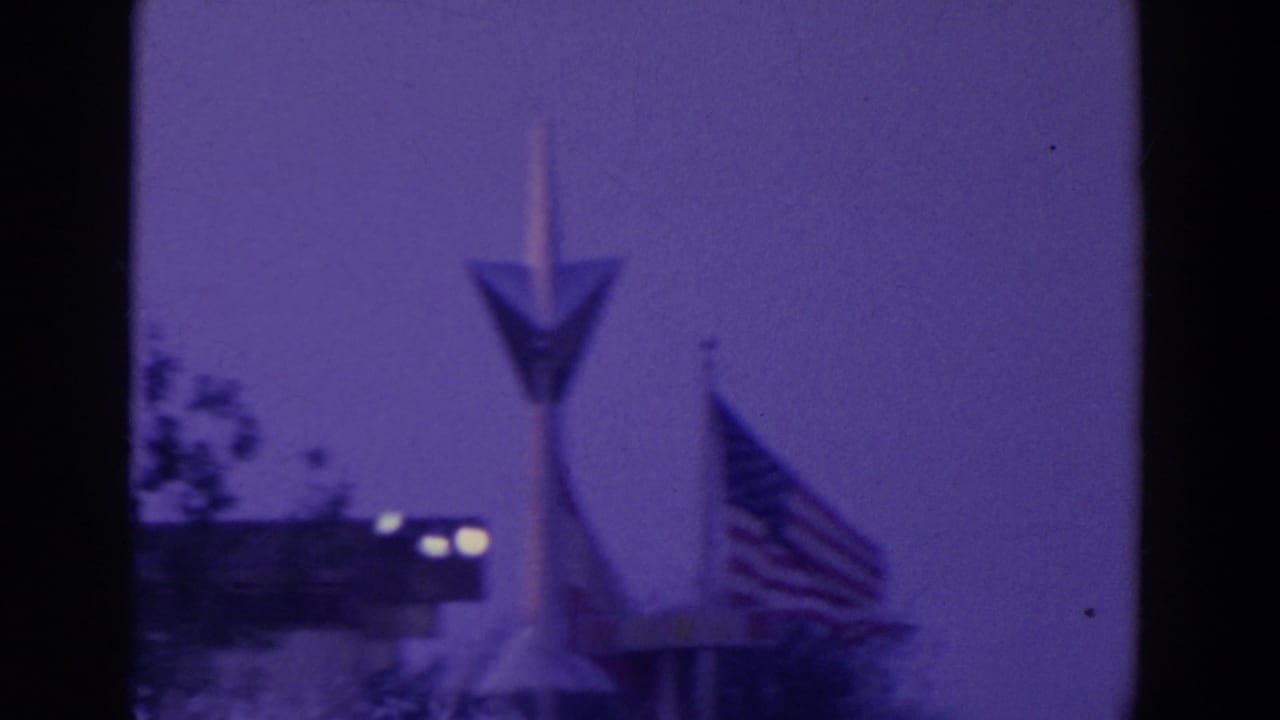 1964 World's Fair home-movie frame showing an arrow-shaped pylon, American flag, and pavilion edge under a purple night sky.
