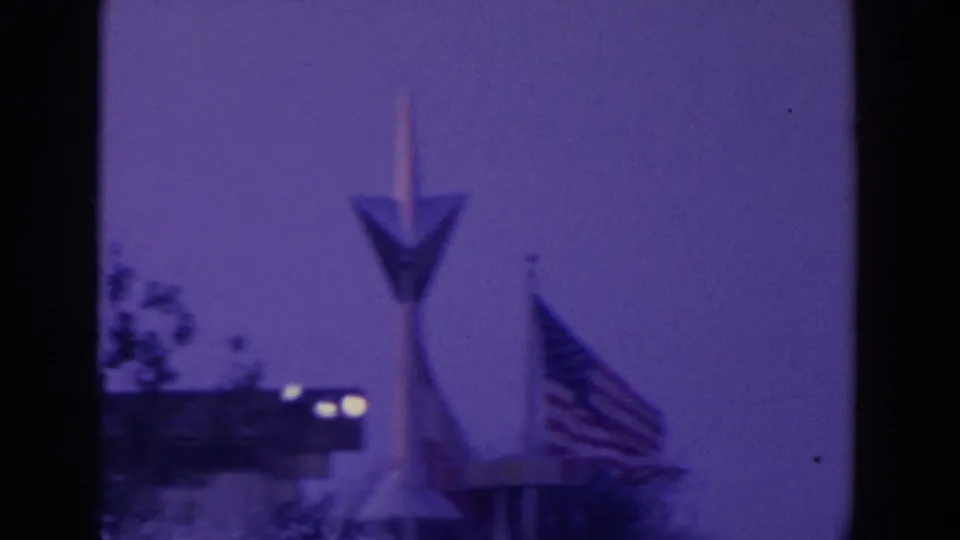 1964 World's Fair home-movie frame showing an arrow-shaped pylon, American flag, and pavilion edge under a purple night sky.