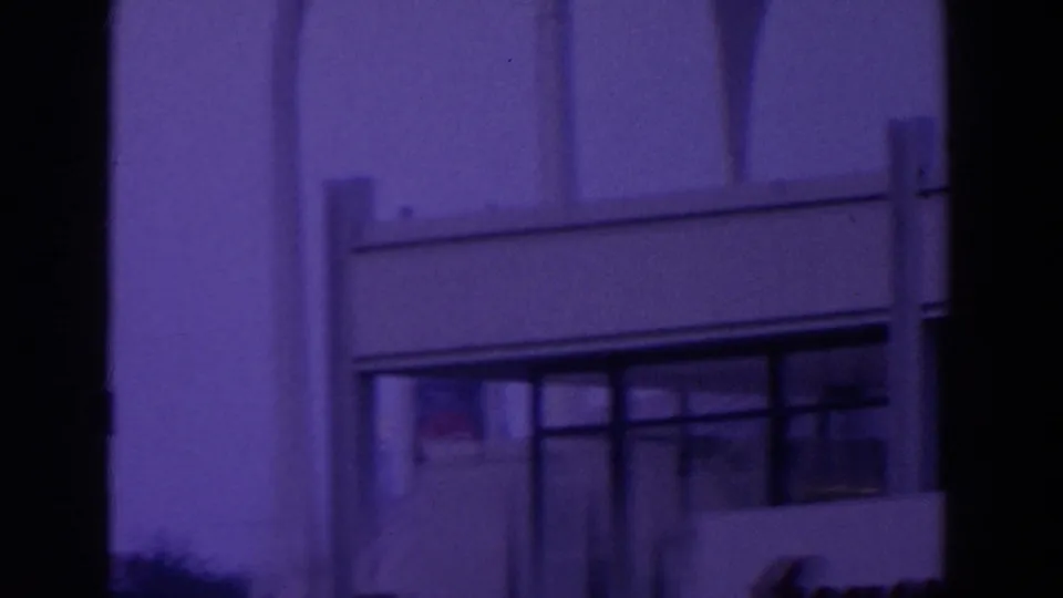 Visitor-shot 8mm frame of a modern fair pavilion facade with pale columns, railing, and dark window bands at night.