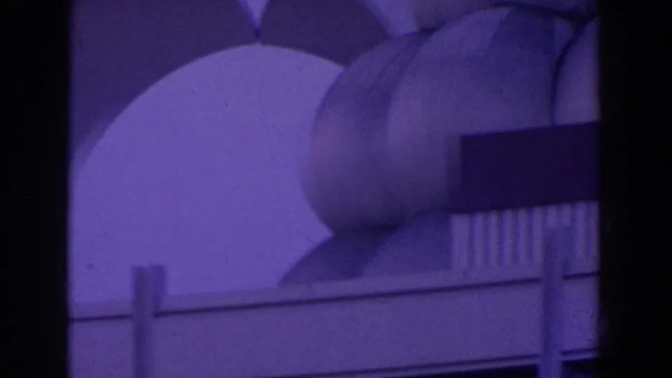 Night fairground frame showing rounded modern pavilion forms, a starburst sign, and dark rectangular bands in purple-tinted 8mm footage.