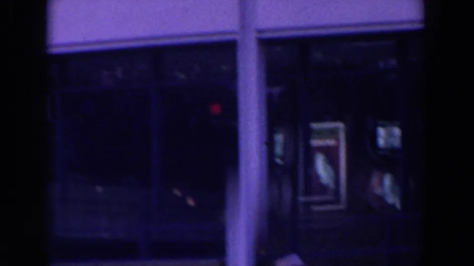 The camera faces a darker building entrance or window bay with poster-like displays and strong purple-blue film coloration.