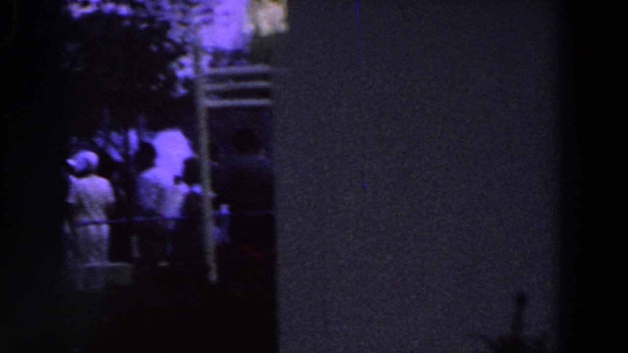 A dim 8mm frame shows people gathered near broad columns and trees, with figures partly obscured by darkness and motion blur.