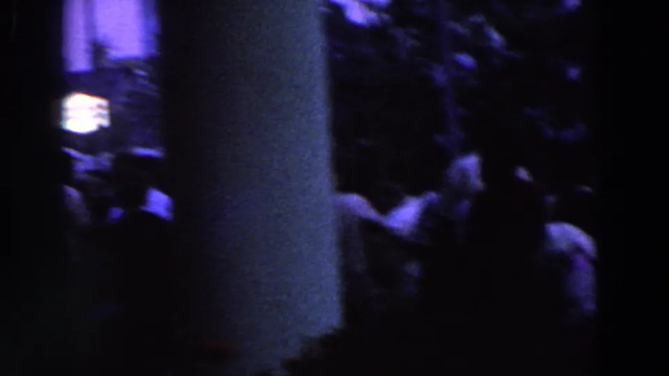 A low-light 8mm view shows figures moving between tall columns, with faces indistinct and the setting mostly in shadow.