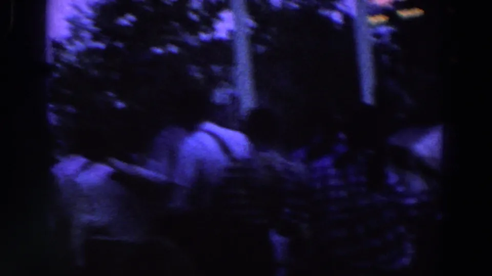 A handheld 8mm frame looks over the backs of people in casual shirts toward trees and columns in a dim gathering area.