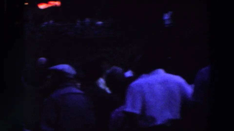 The frame shows a dark crowd scene with several people grouped together and one light-shirted figure prominent in the foreground.