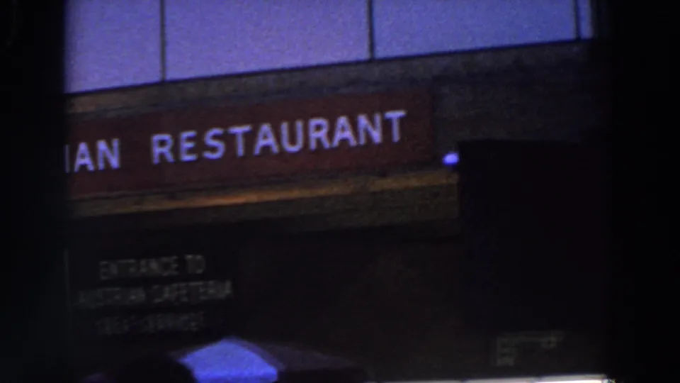 A closer 8mm view of the Austrian Restaurant sign shows partial lettering, a dark doorway, and blurred people in the foreground.