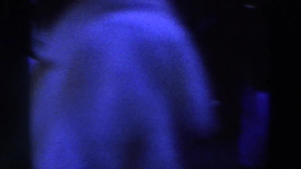Underexposed 8mm frame with a blue-lit partial figure close to the camera and surrounding darkness.