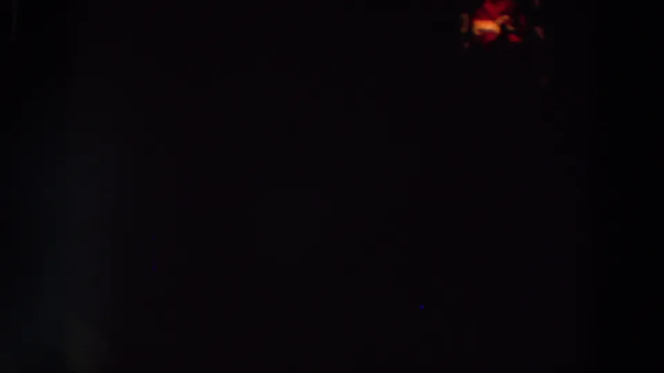 Very dark 8mm night frame showing a tiny orange light point and a faint vertical blue flare or scan artifact against the black field.