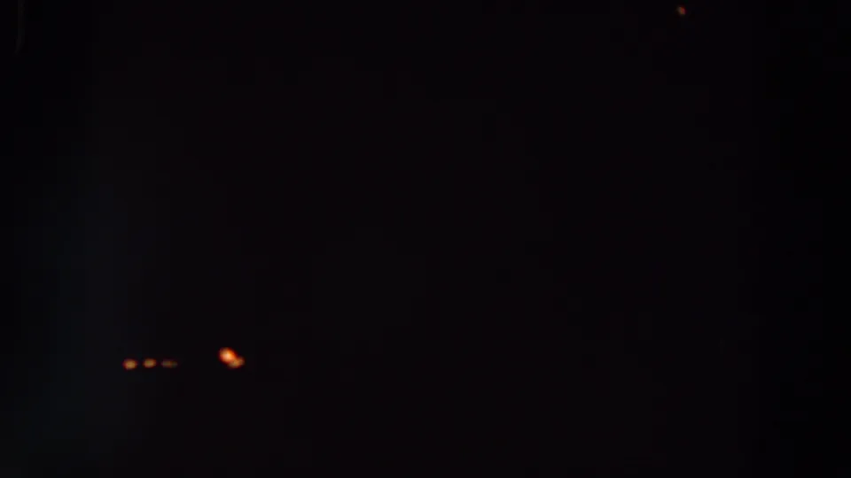 Low-light 8mm frame with a cluster of small orange points near the lower left, surrounded by heavy shadow and visible film grain.
