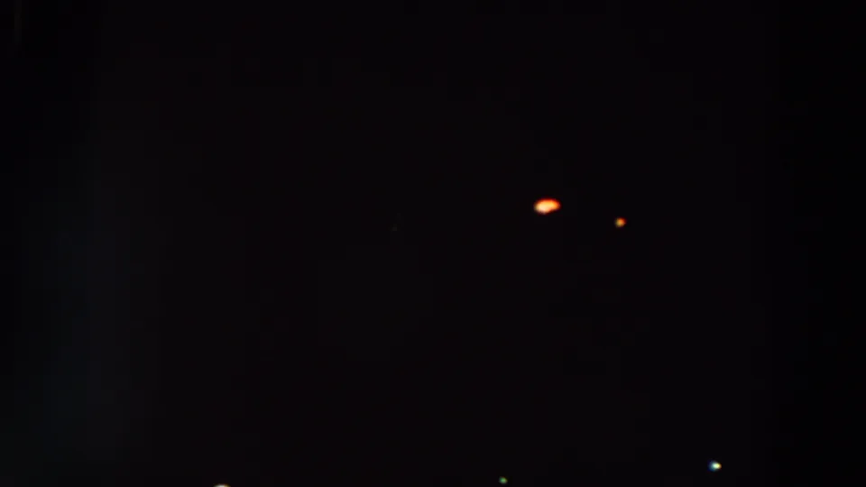 Underexposed 8mm night view with small orange, green, and blue light points scattered across the lower portion of the frame.