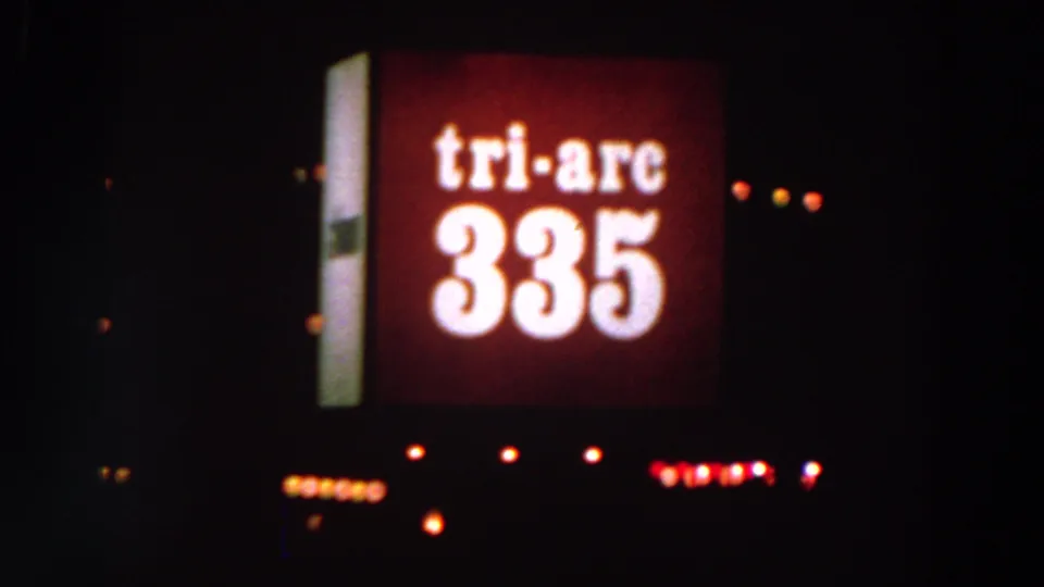 Nighttime 8mm frame showing a red Tri-Arc 335 sign against a dark fairground or urban setting, with small lights below.