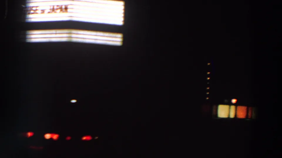 Nighttime frame with bright horizontal panels at left, scattered red lights below, and dark building shapes in the background.