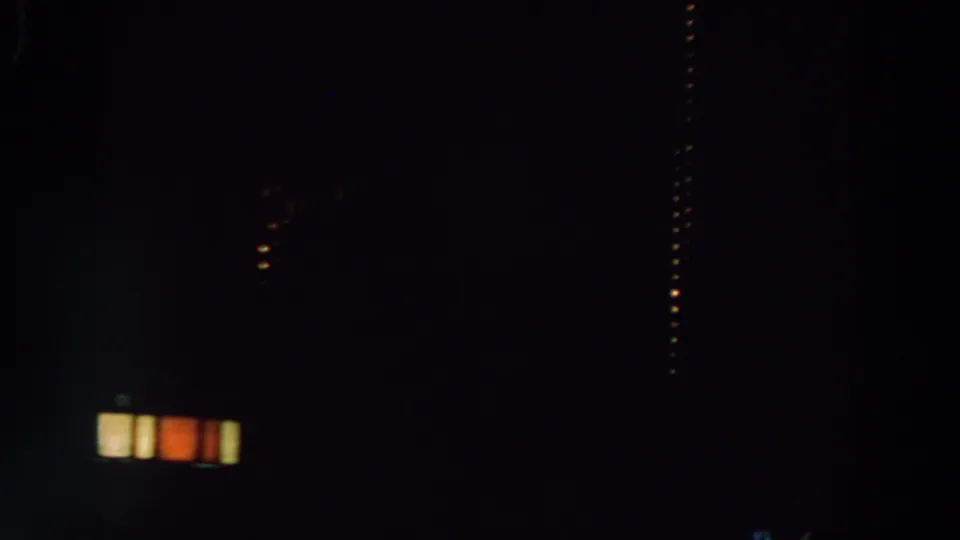 A dim 8mm nighttime frame shows a dark structure with small illuminated windows and scattered lights in the distance.