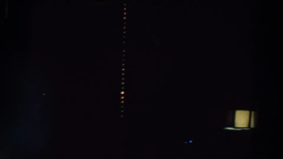 Dark 8mm frame from the reported 1964 World's Fair footage showing faint architecture and a narrow vertical line of warm lights.