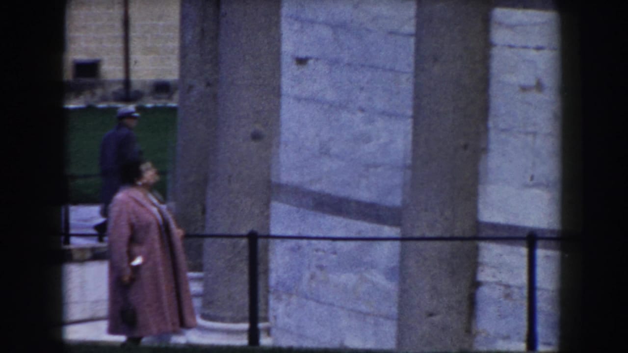 Woman in a pink coat walks beside stone columns and a low railing.