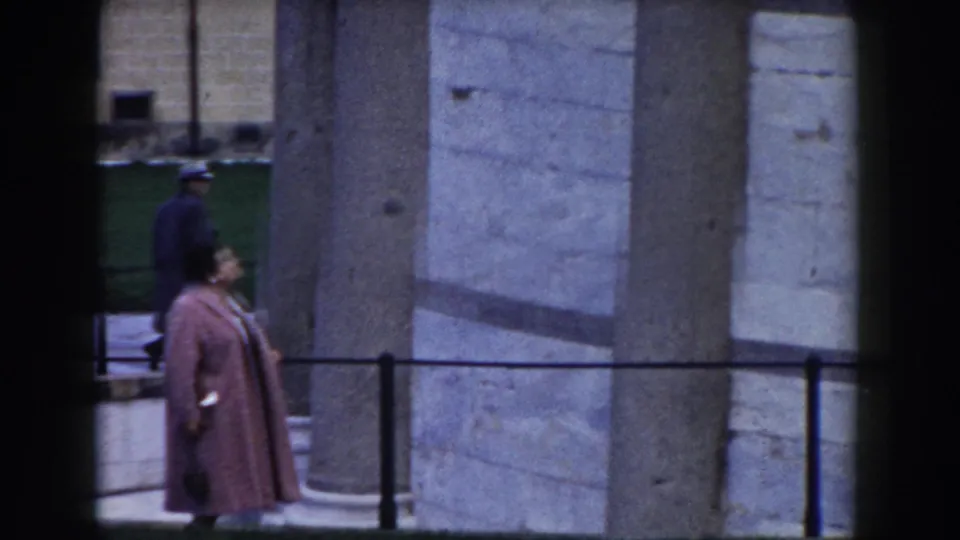 In Pisa, Italy, a woman in a long pink coat and dark hat walks near the Leaning Tower's stone columns in 1959 home-movie footage.