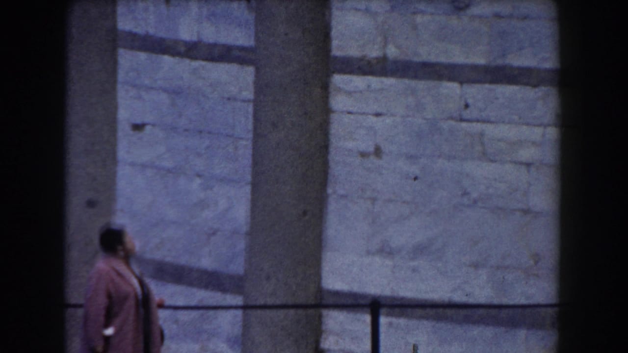 Woman in a pink coat looks upward beside pale stone masonry.