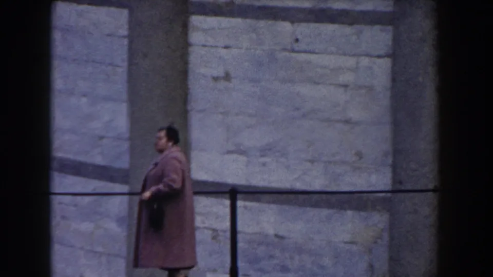 In Pisa, Italy in 1959, a woman in a pink coat carries a dark handbag while walking past tan stone columns and pale masonry.