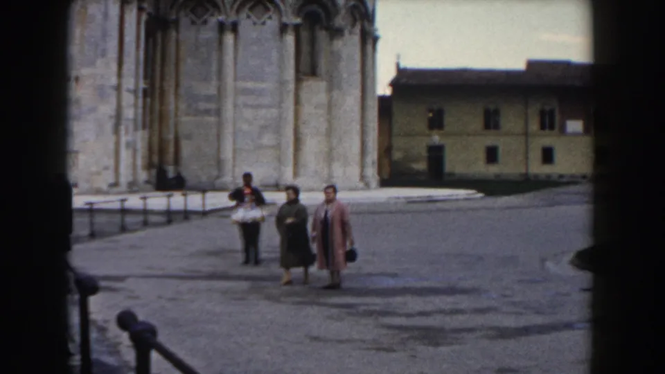 In Pisa, Italy, two women in mid-century coats walk hand-in-hand near cathedral stonework while another pedestrian crosses nearby.