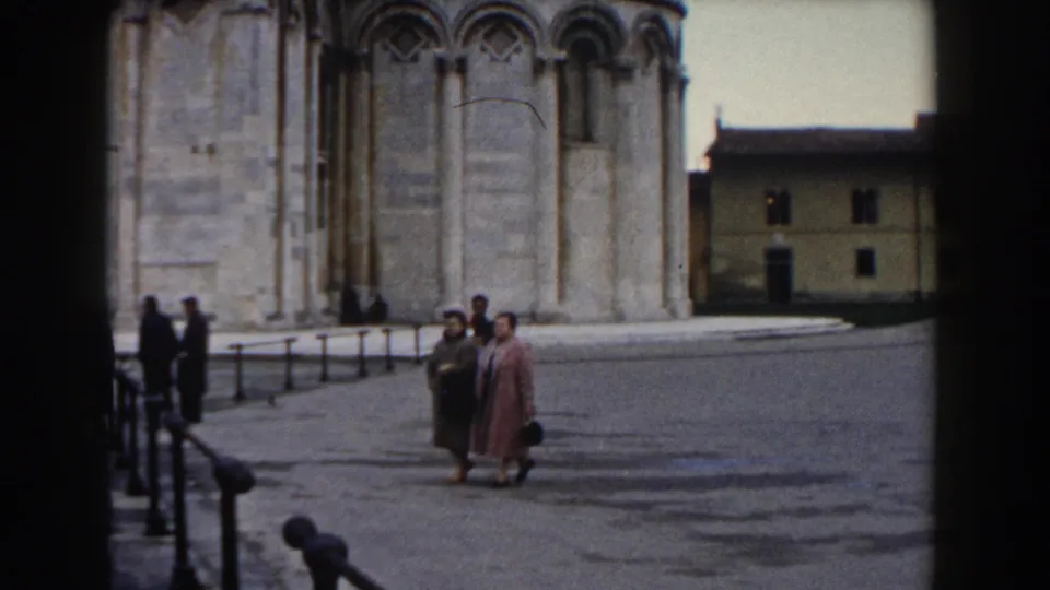 Two women in coats move through the Pisa piazza, with pedestrians gathered near the cathedral wall and railings.