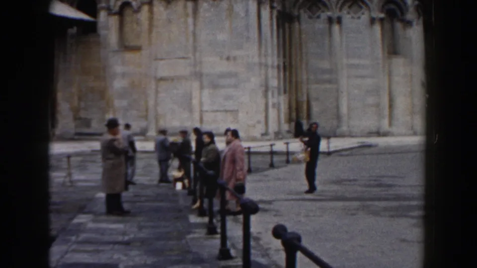 Visitors in mid-century clothing move and pause near the Pisa cathedral facade, with the home-movie frame showing railings and piazza paving.