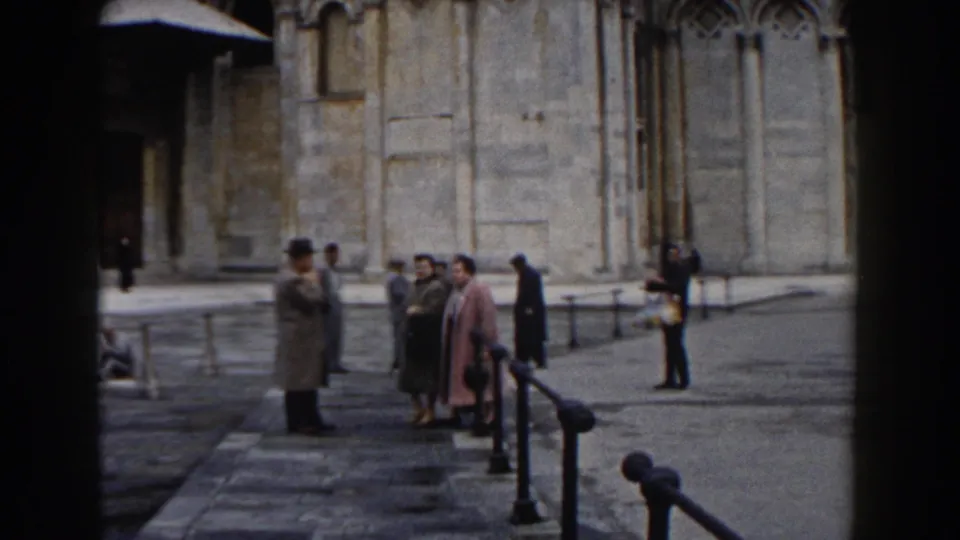 1959 Pisa travel footage shows tourists in coats and hats standing near tan stone architecture in a paved courtyard.
