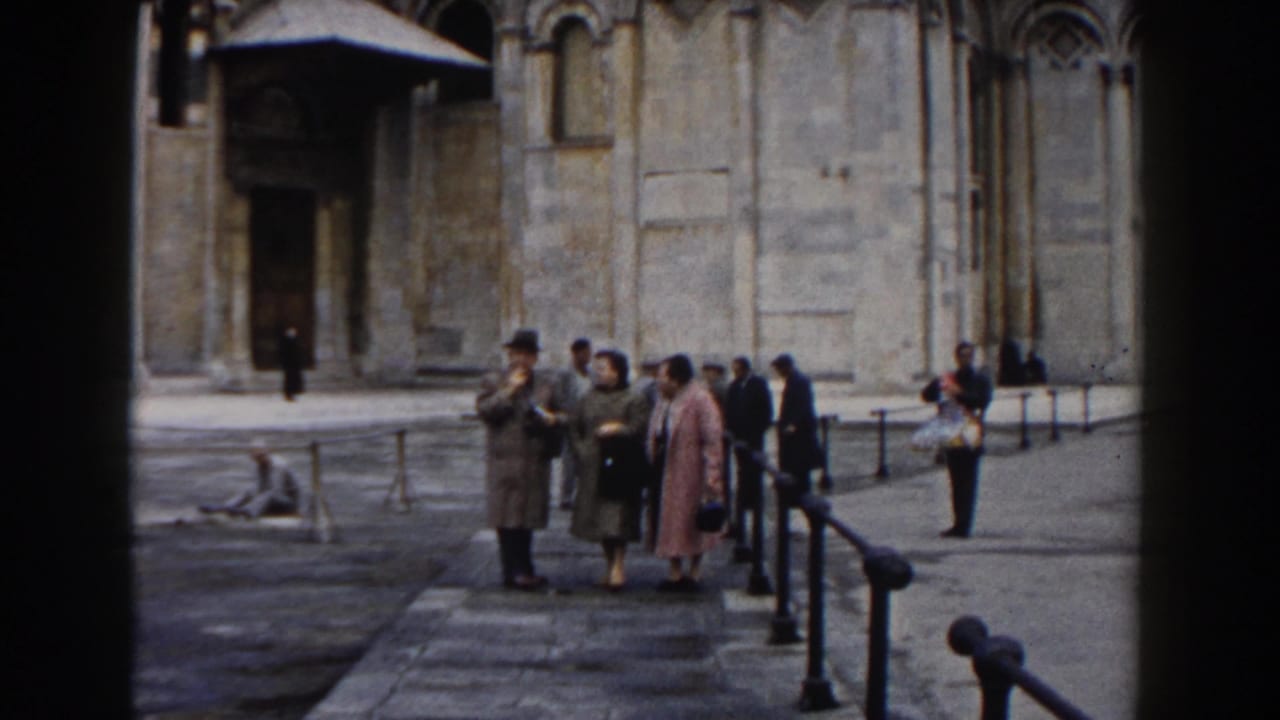 Tourists gather in front of a tan stone building with railings and arched details.
