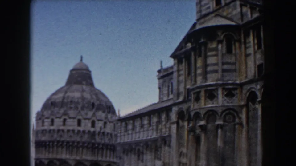1959 Pisa travel-film view with the baptistery dome and cathedral side facade framed against a clear blue sky.