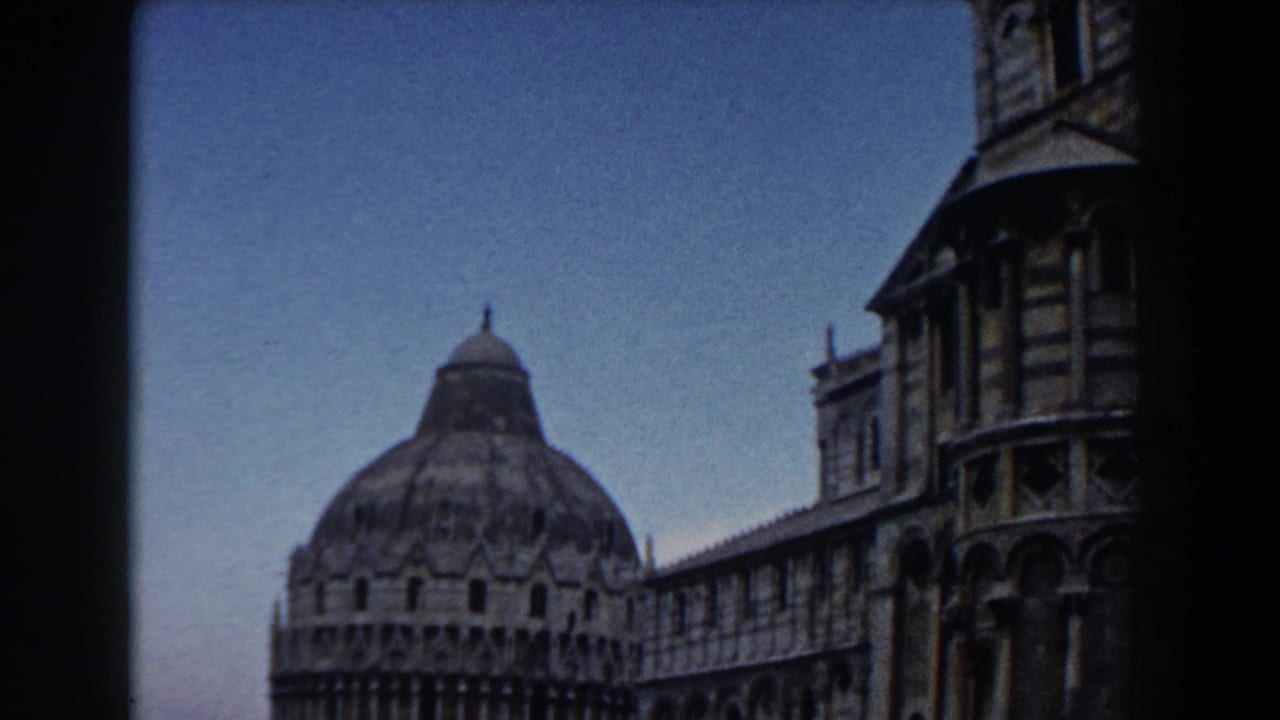 1959 8mm view of Pisa's Baptistery with part of the Cathedral at right, framed across the grassy piazza under a pale sky.
