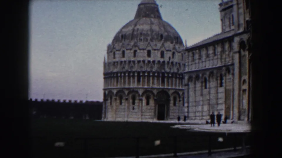 Wider 1959 8mm view in Pisa showing the Baptistery and Cathedral beside a lawn, with distant visitors on the walkway.
