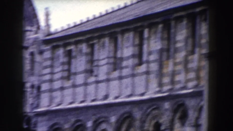Handheld 1959 Pisa footage frames a long stone facade and arched lower level partly blocked by a dark foreground shape.