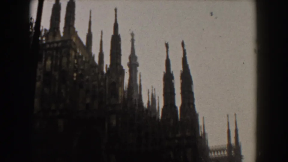 1950s Italian travel footage frame with Gothic cathedral spires, roof details, and dark window borders in soft focus.
