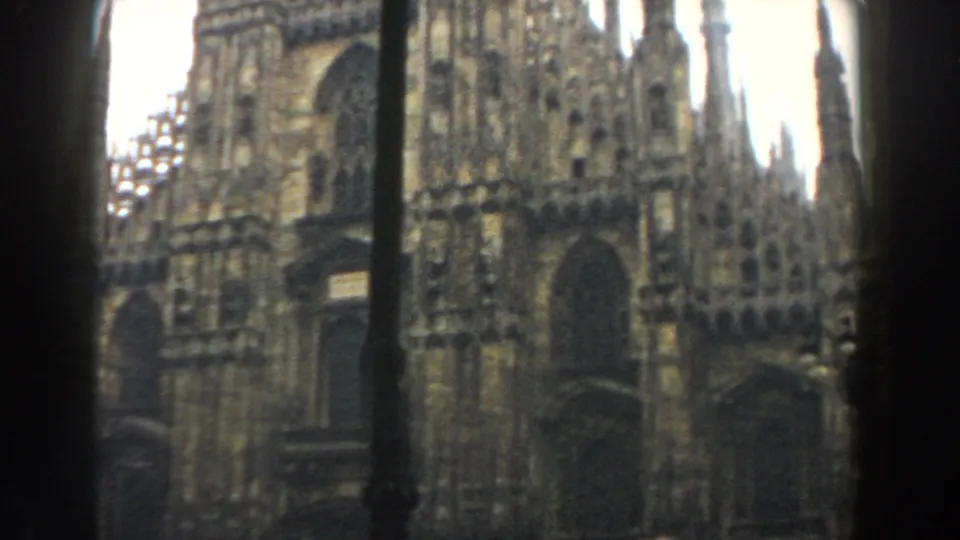1959 8mm frame showing the cathedral's arched facade and spires, likely Milan's Duomo, with a dark foreground pole and soft exposure.