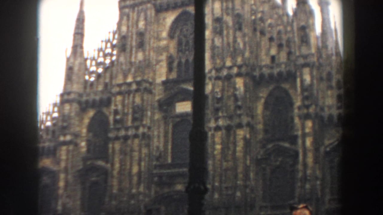1959 8mm travel view of Milan Cathedral's ornate Gothic facade, seen through dark foreground framing under an overcast sky.