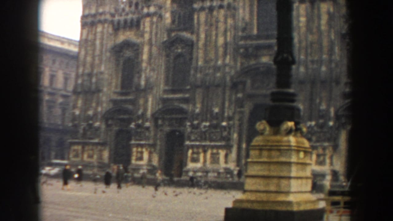 Pedestrians cross a paved piazza before an ornate stone facade and lamp posts.