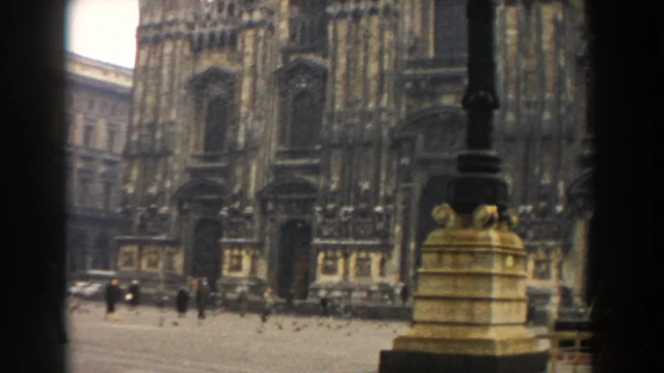 1959 Pisa home-movie frame showing scattered pedestrians in a paved square before ornate tan stone architecture.