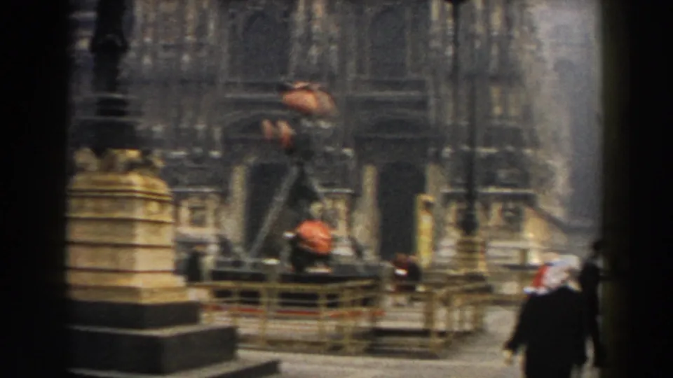 1959 Pisa frame with a close view of a stone monument, dark lamp posts, and an ornate cathedral entrance beyond.