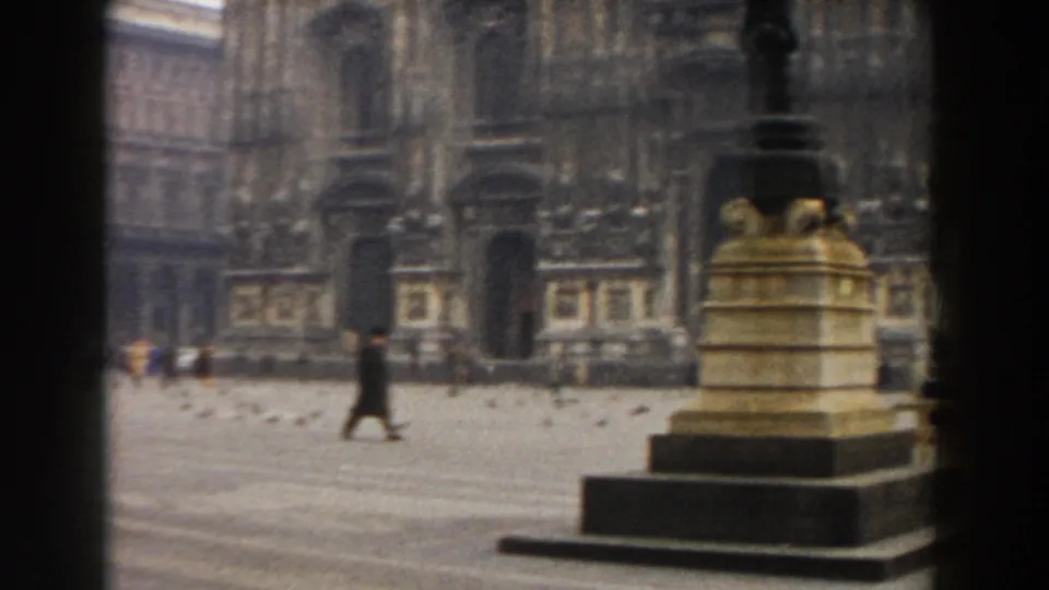 1959 Pisa home-movie frame showing a stone monument in the foreground and pedestrians crossing the open square.