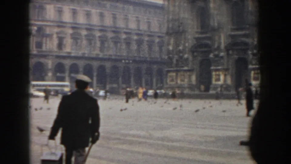 1959 Pisa frame showing a uniformed person in the foreground, pigeons on the pavement, and tan stone buildings across the square.