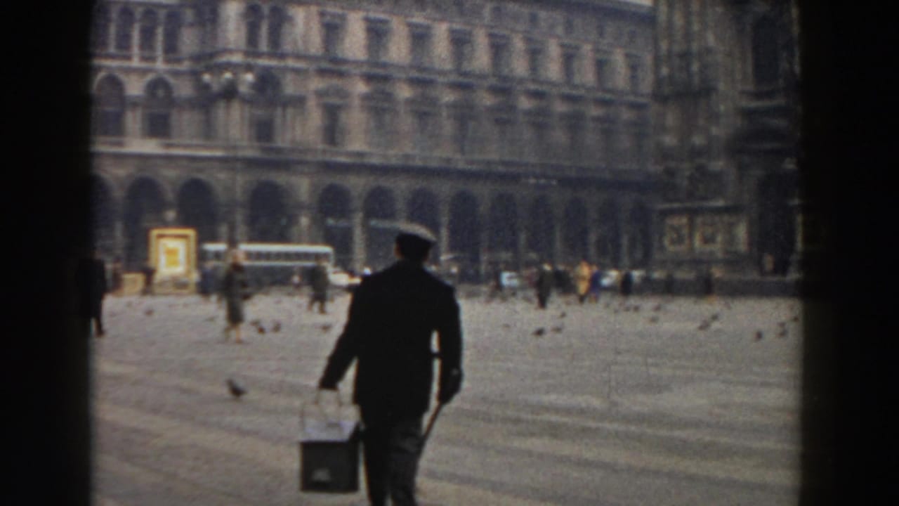 Man in dark jacket and cap carries a bucket across a large square with pigeons and arched buildings behind him.