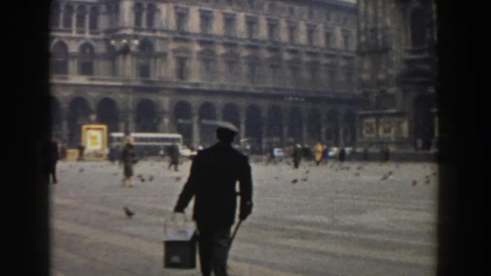 1959 Pisa home-movie frame showing a man in dark clothing carrying a bucket through a broad historic square with pigeons, pedestrians, and buses in the distance.