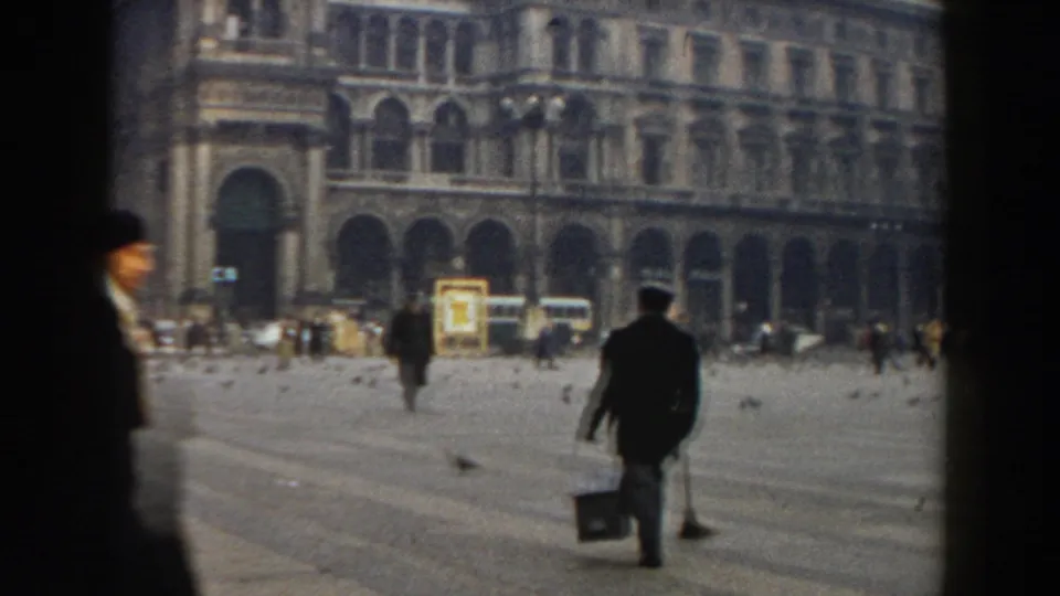 1959 Pisa frame with a dark-clad man walking away across the open square, carrying a bucket as pedestrians, pigeons, and buses appear near historic arcades.