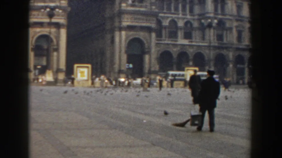1959 Pisa home-movie view of two dark-clad figures in a broad square, one carrying a bucket, with pigeons scattered before arched historic buildings.