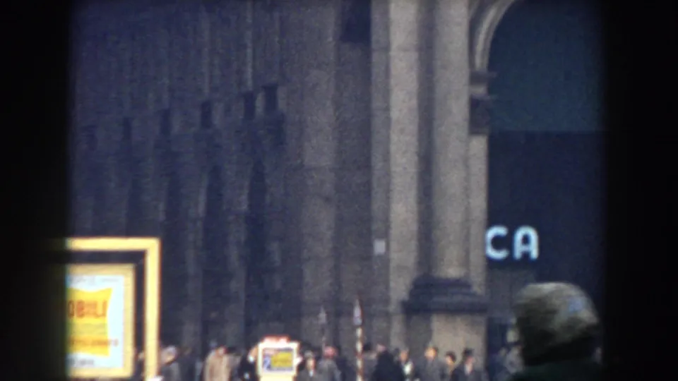 1959 Pisa street view with pedestrians near stone columns, a bright yellow sign, and partial blue lettering at the right edge.