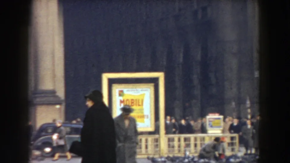 1959 Pisa frame showing a woman in a dark coat passing a yellow street sign, with pedestrians, barriers, and cars behind her.